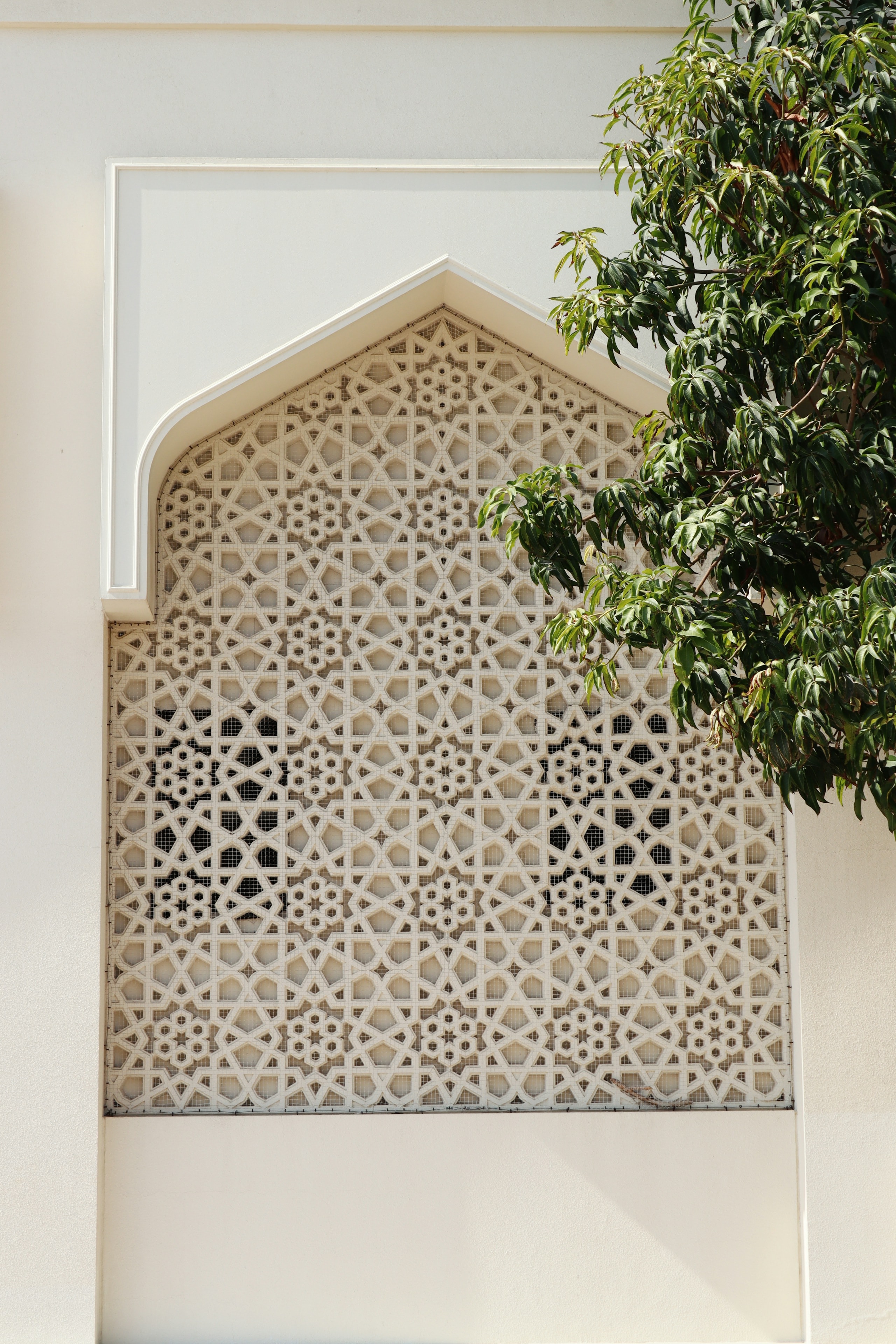 A white carved lattice window in a pointed arch beside a tree